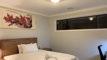 A neatly made bed with white linens, a wooden headboard, and a rolled towel on the bed. There is a nightstand with a lamp and a tissue box beside the bed. A large painting of red flowers hangs on the wall above the bed.