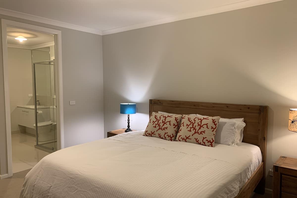 Une chambre à coucher avec un grand lit, une tête de lit en bois, une table de chevet avec une lampe, et une salle de bain visible à travers une porte en verre.