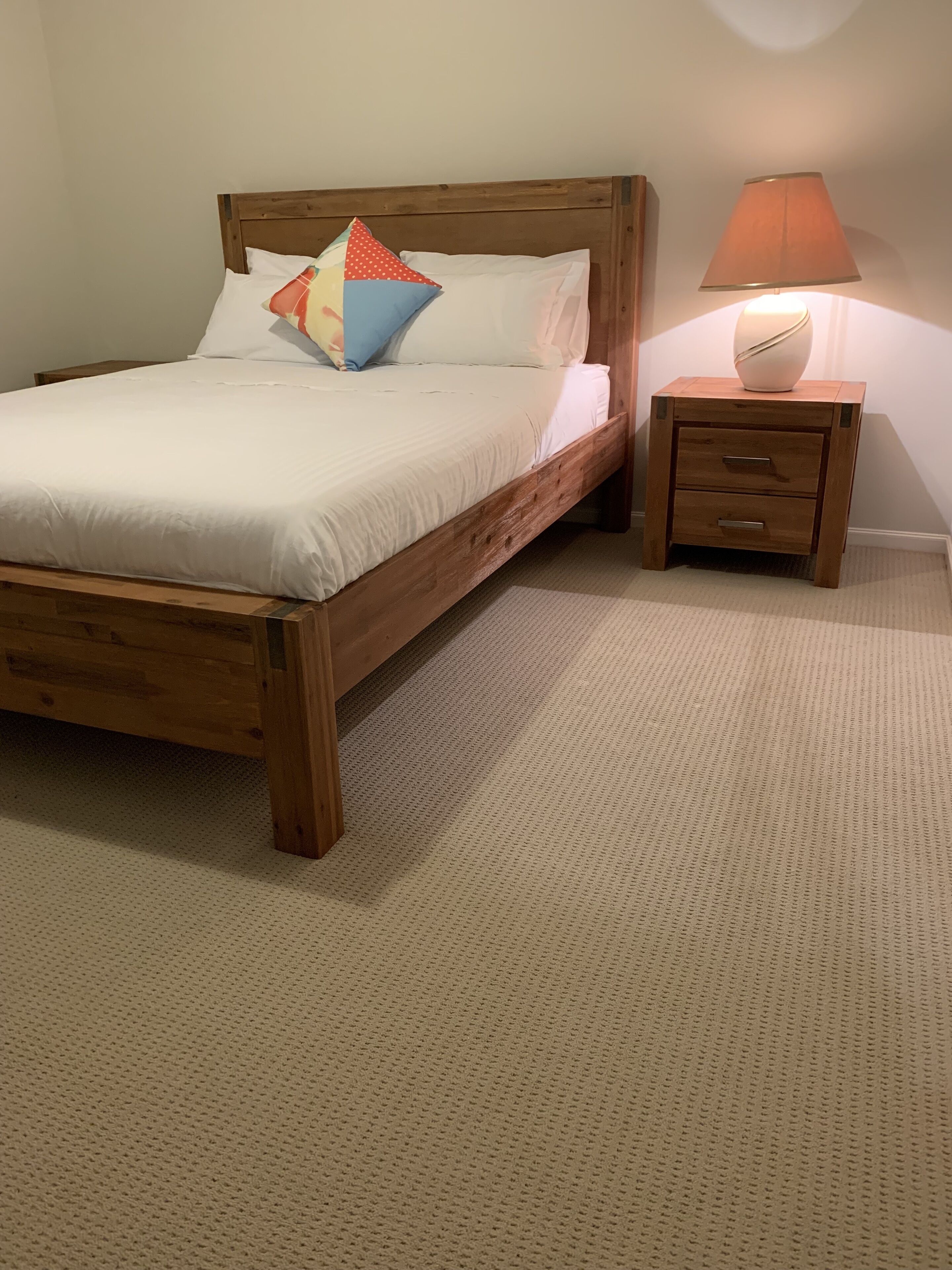 Ein hölzernes Bett mit weißer Bettwäsche und bunten Kissen, ein kleiner Nachttisch mit Lampe sowie ein strukturierter, beigefarbener Teppich.