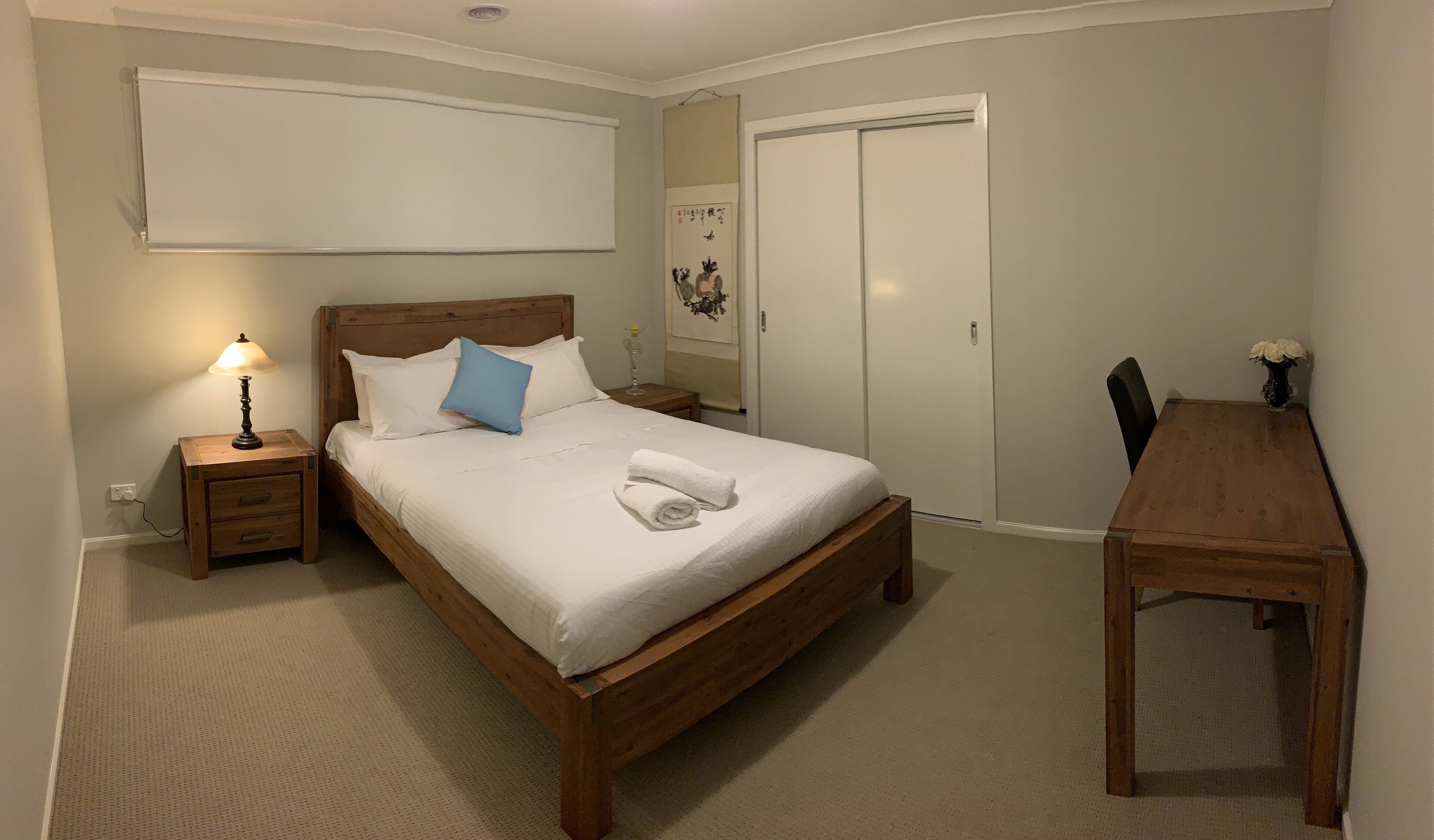 Ein ordentlich bezogenes Bett mit weißen Leinentüchern und einem blauen Kopfkissen, ein Nachttisch mit Lampe, ein Holztisch und ein weißer Kleiderschrank in einem schlichten, aufgeräumten Zimmer.