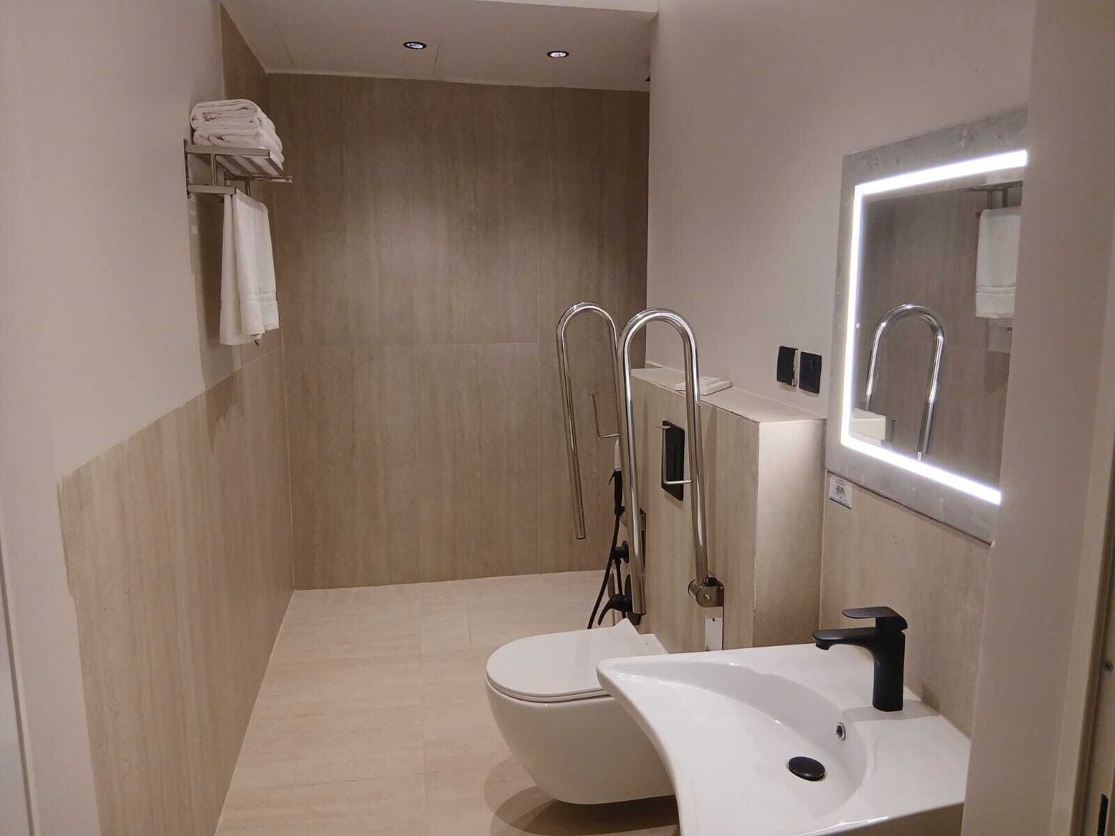 Un baño moderno con inodoro blanco, lavamanos con grifo negro, espejo con iluminación integrada y toallero con toallas dobladas.