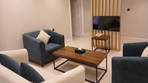 A modern living room with a blue sofa, a white armchair, a wooden coffee table, and a television on a wooden stand. - Raoum Inn Serviced Apartments-Shaqraa (Shaqra)