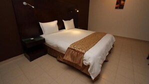 A neatly made bed with a white and brown bedspread, a bedside table with a lamp, and a wall-mounted light fixture. - Raoum Inn Serviced Apartments-Majmaa (Al Majmaah)