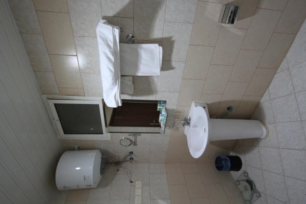 Un baño pequeño con inodoro, lavamanos y ducha. Hay un perchero sobre el inodoro y una pequeña repisa con artículos de aseo.