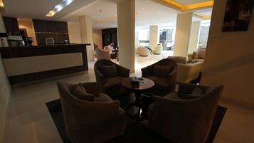 A modern hotel lobby with a reception desk, seating area with sofas, and a large open space with a dining area.