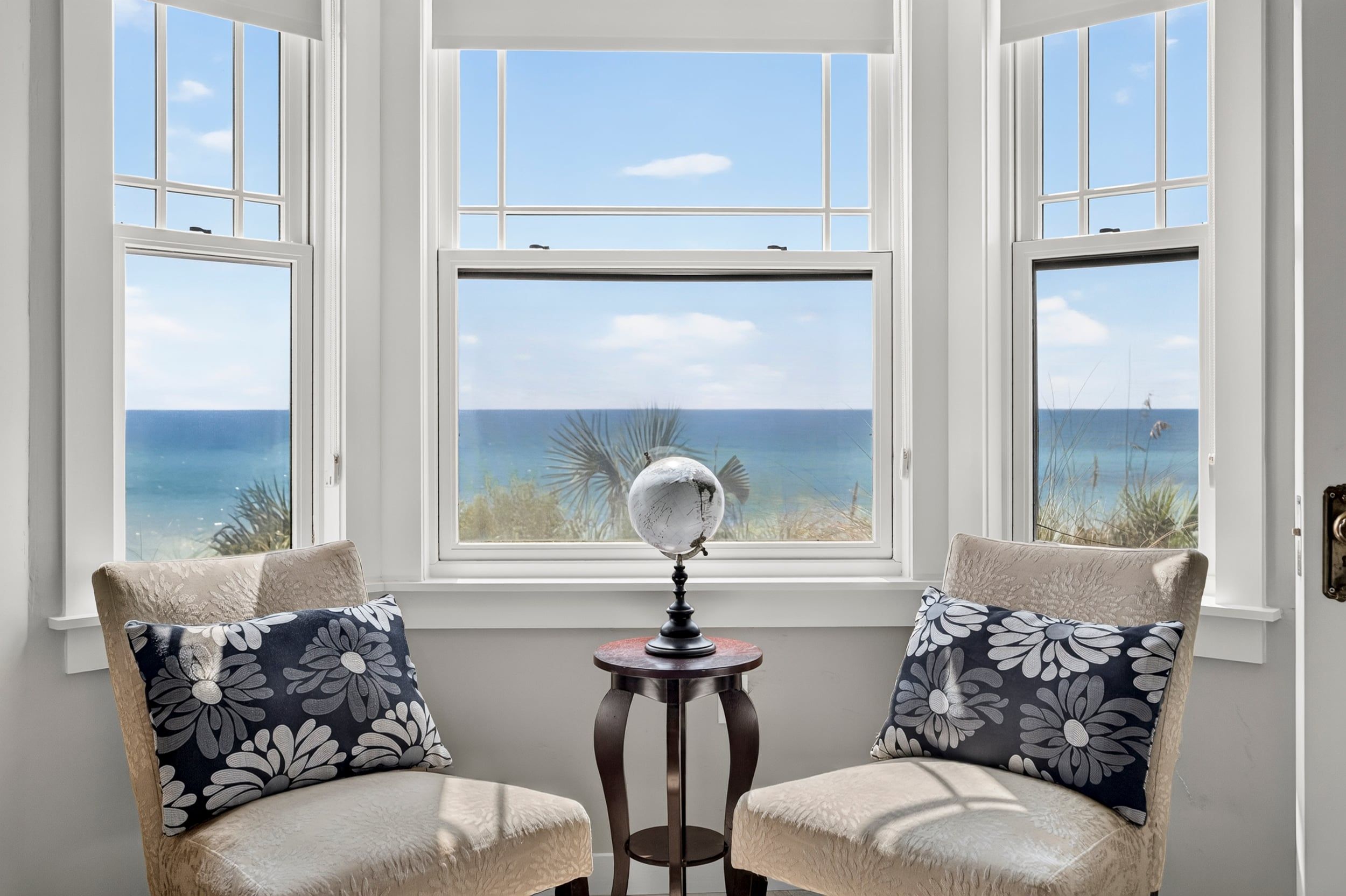 Una habitación con vistas al mar a través de tres ventanas, dos sillones con cojines estampados y un globo terráqueo sobre una mesita.