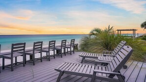 Een houten terras met lounge stoelen met uitzicht op een strand bij zonsondergang.