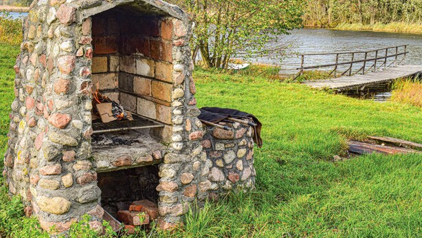 Un four extérieur en pierre, avec un feu qui crépite à l’intérieur, entouré d’herbe et bordé d’une jetée en bois donnant sur une étendue d’eau.