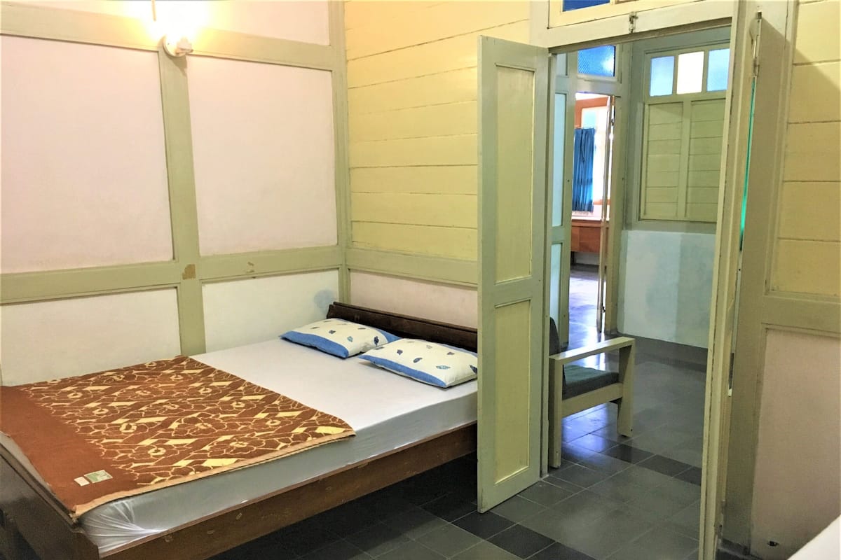 A small, single-bed room with a wooden bed frame, a patterned bedspread, and a blue pillow. The room has a tiled floor and a door leading to another room.