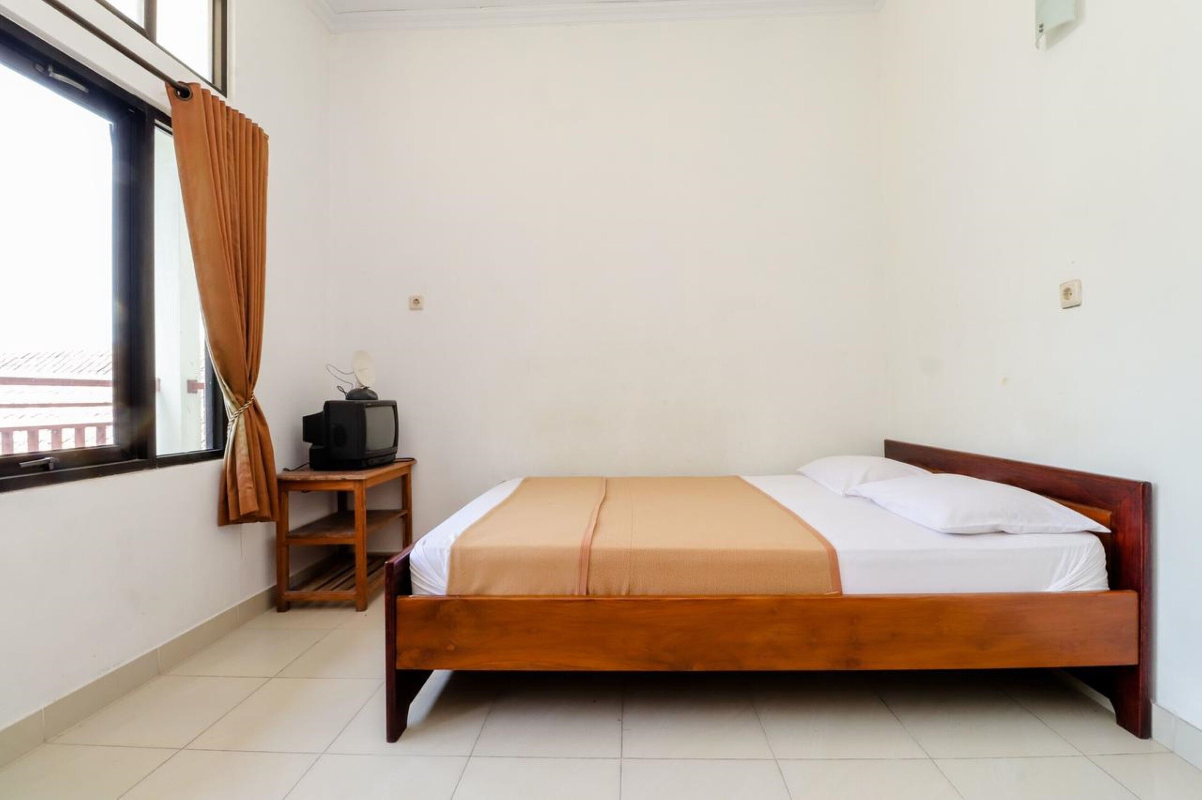 A bedroom with a wooden bed, a small wooden nightstand with a television, and a window with curtains.