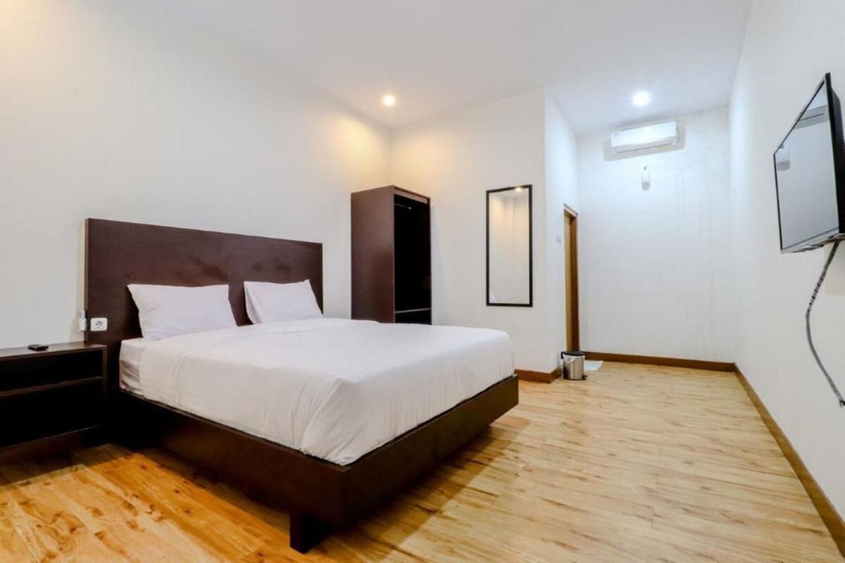 A bedroom with a wooden floor, a bed with white linens, a dark headboard, a nightstand, a wardrobe, a mirror, and a TV mounted on the wall.