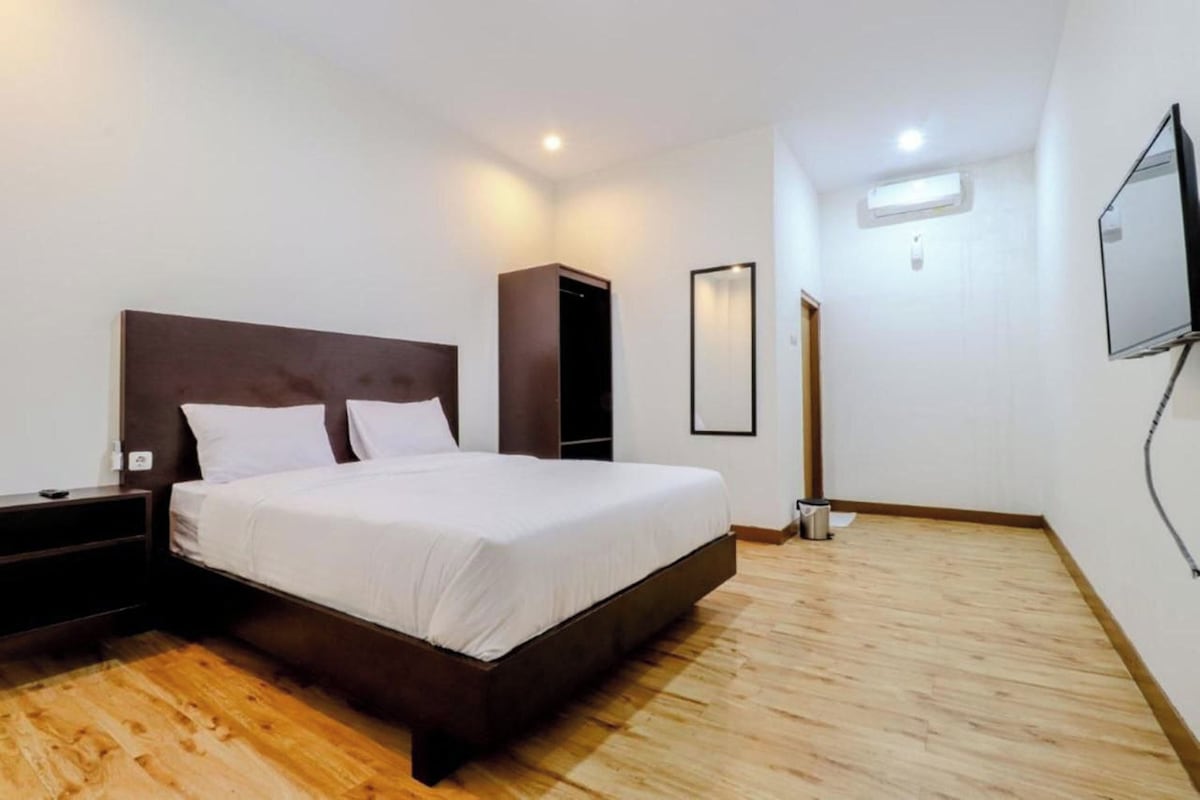 A bedroom with a wooden floor, a bed with white linens, a dark headboard, a nightstand, a wardrobe, a mirror, and a TV mounted on the wall.