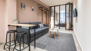 A modern living room with a sofa, two bar stools, a small table, and a TV mounted on the wall. - Filia Place (Athens)