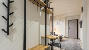 A modern hotel room with a wooden bench, a small table, a chair, and a desk. There is a plant on a shelf, a door, and a wall-mounted shelf with a mirror. - Filia Place (Athens)