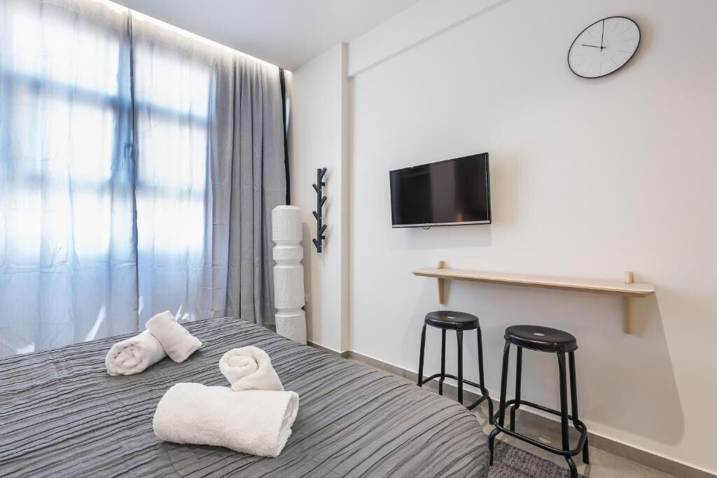 A hotel room with a bed, a TV mounted on the wall, a clock, and two bar stools.