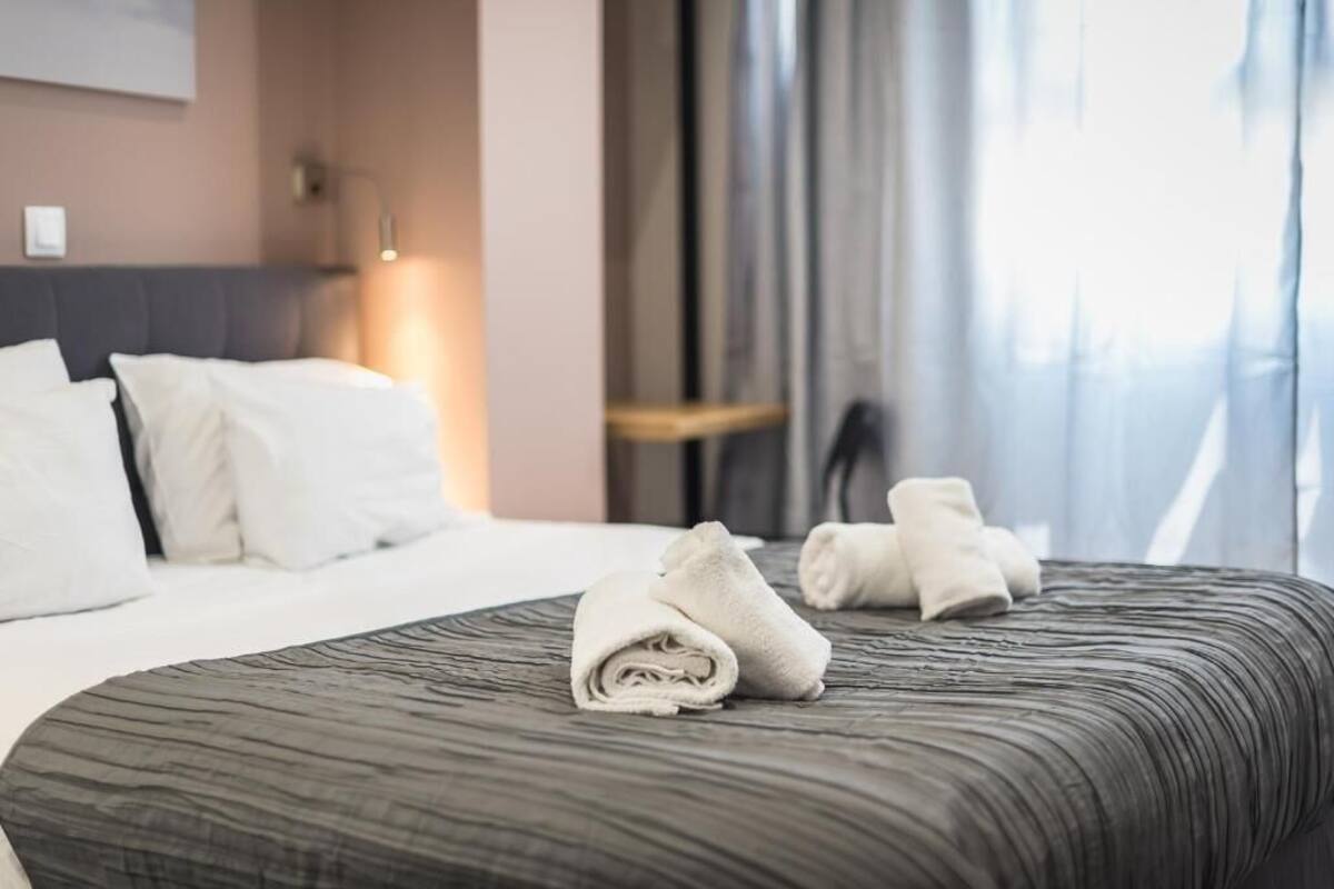 A neatly made bed with a grey textured bedspread and two rolled towels on it.