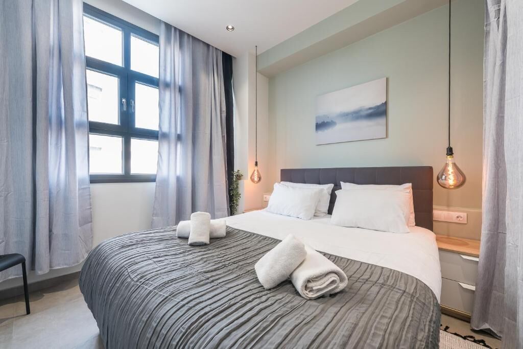 A modern hotel room with a large bed, grey bedding, a view of the city, and a painting on the wall.