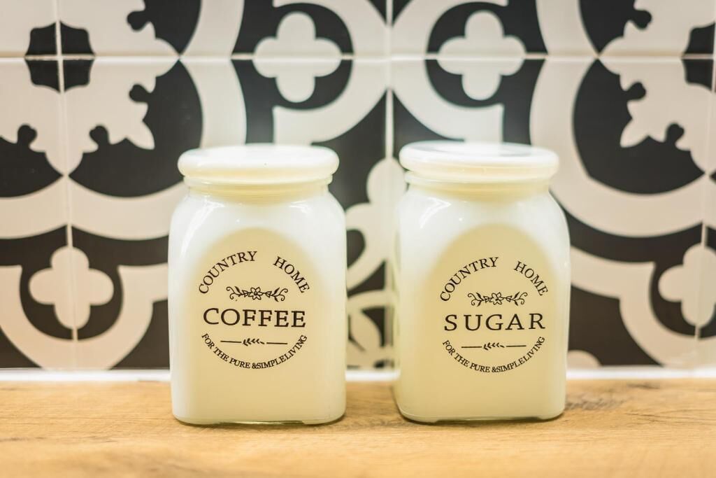 Twee glazen potten met de tekst 'Country Home Coffee' en 'Country Home Sugar' op een houten oppervlak, met een betegelde muur met patroon op de achtergrond.