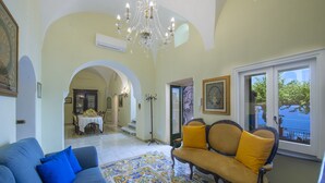 Un salon spacieux avec un canapé bleu, un fauteuil jaune et un tapis à motifs. On y trouve un lustre, un coin repas à l’arrière-plan et une vue à travers une grande fenêtre.