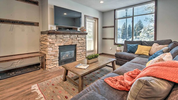 A living room with a stone fireplace, a television mounted above it, a large window with a view of trees, and a gray sofa with colorful pillows. - Solitude Mountain Slopeside Ski Condo w/ Hot Tub! (Brighton)