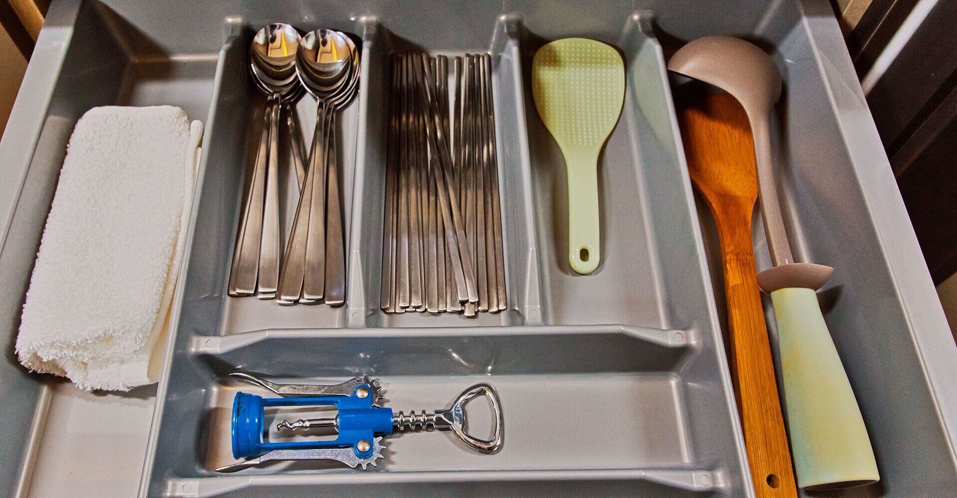 A drawer containing kitchen utensils including spoons, forks, a brush, a spatula, and a bottle opener.