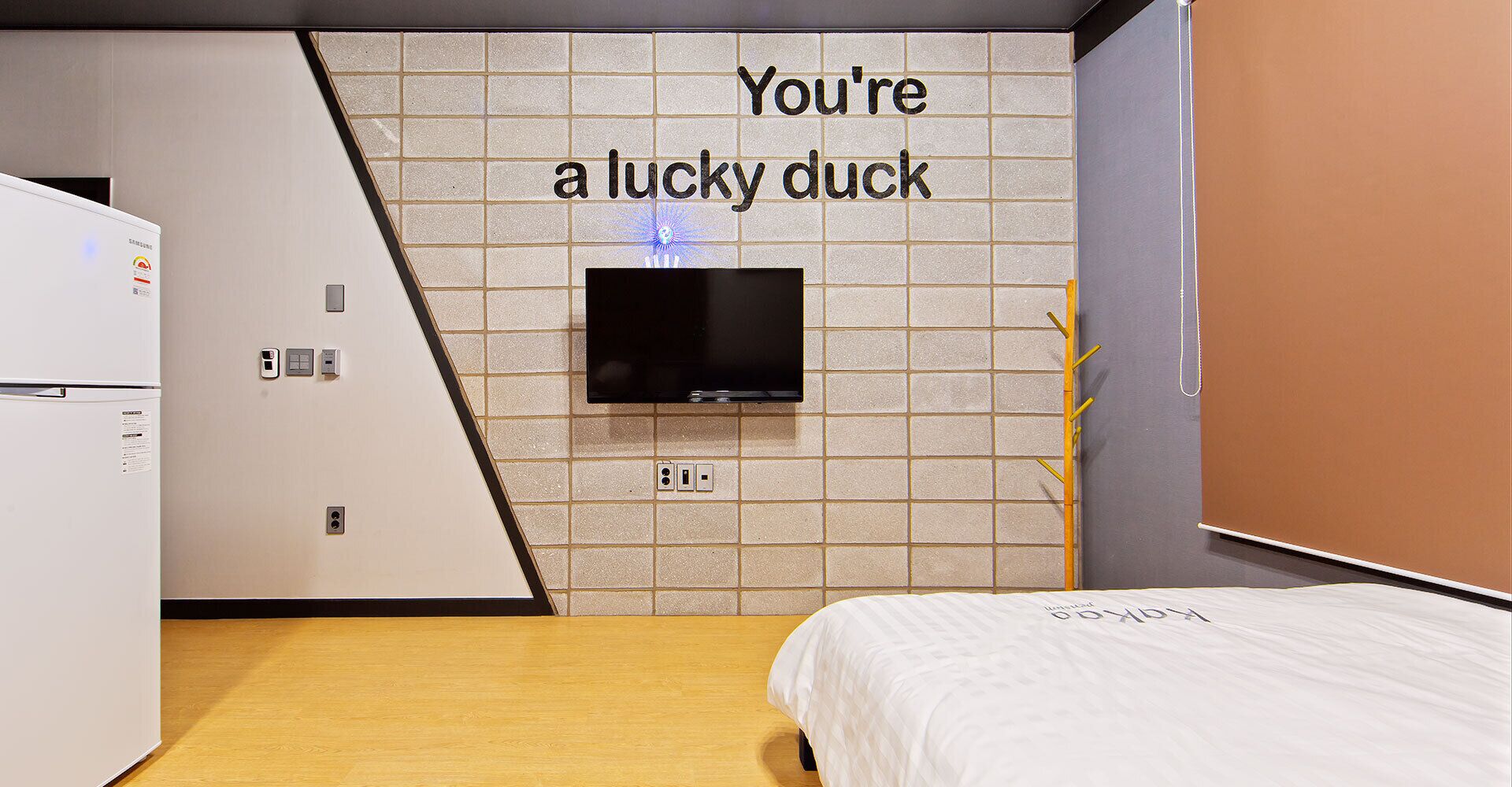 A modern hotel room with a bed, a TV mounted on a tiled wall, and a refrigerator.