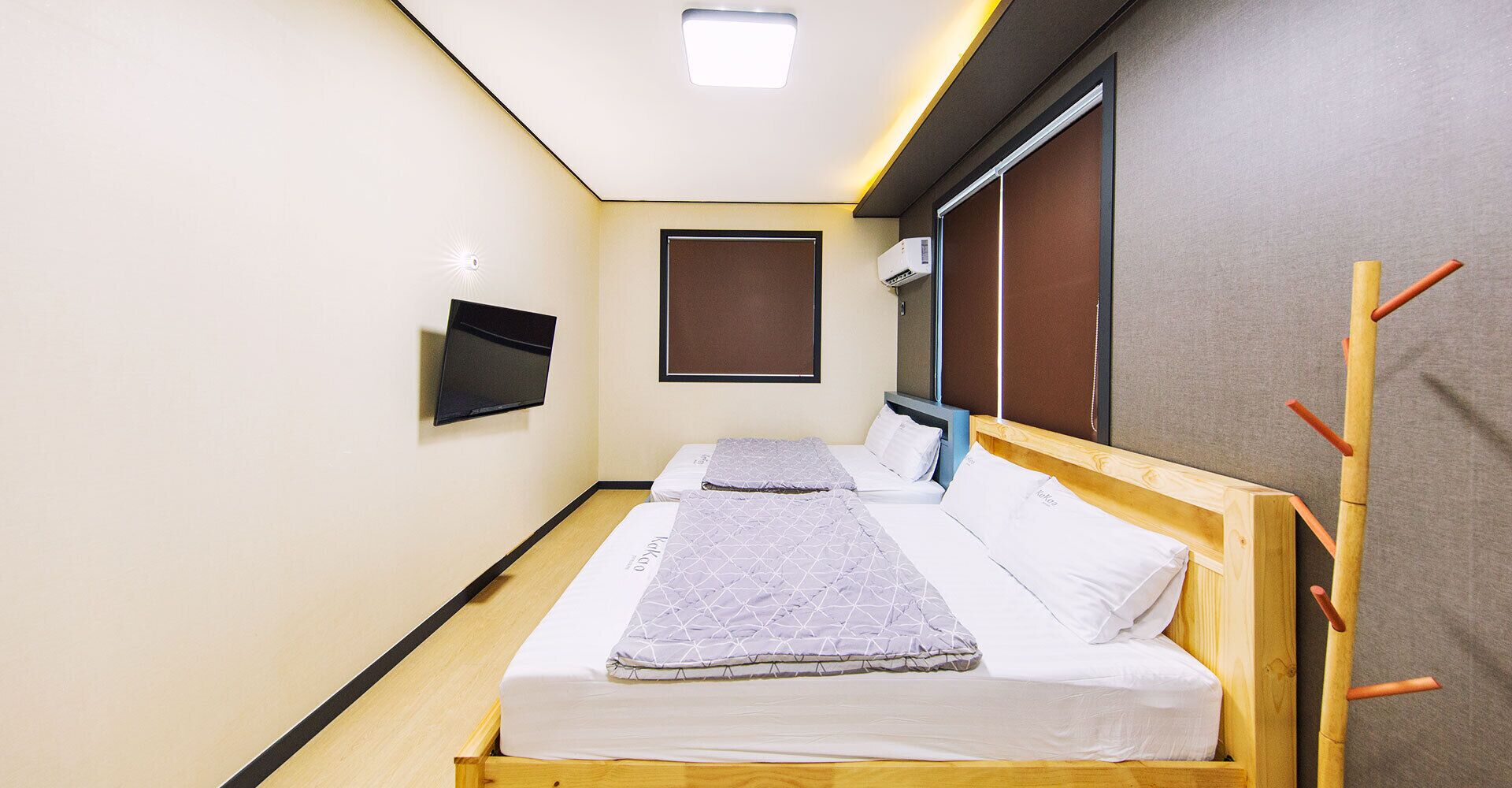A small, modern hotel room with a single bed, a flat-screen TV mounted on the wall, and a window with a brown blind.