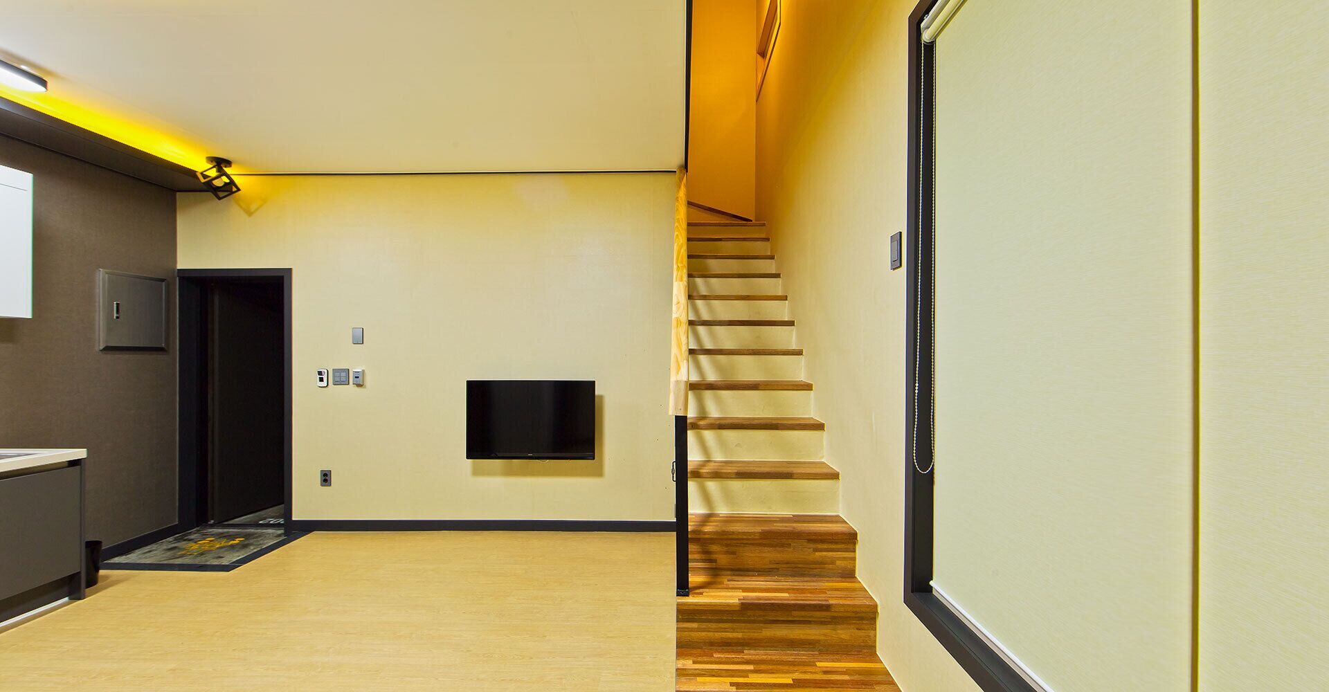 Un couloir d’hôtel moderne avec un escalier en bois, un téléviseur à écran plat fixé au mur et une porte dotée d’un interrupteur.