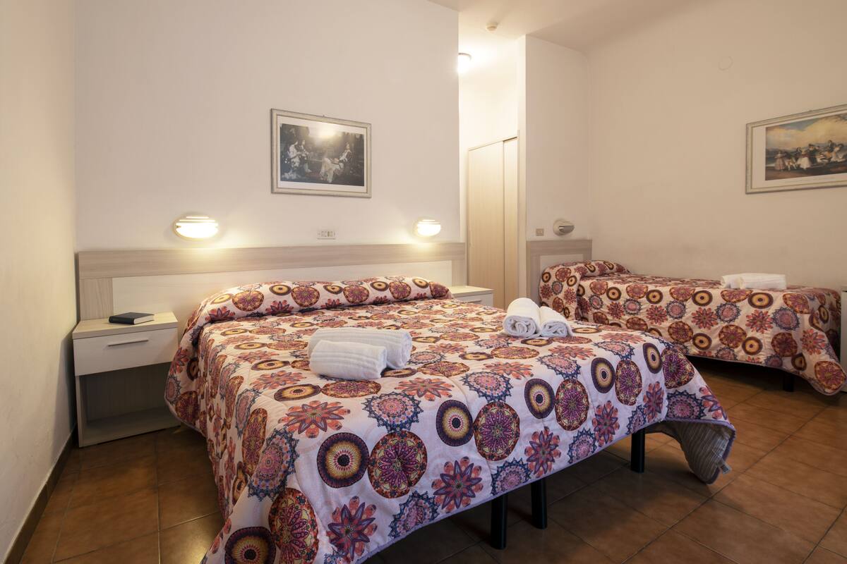 Ein Hotelzimmer mit zwei Betten, jeweils mit gemusterten Bettbezügen, einem Nachttisch mit einem Buch und einer Wandleuchte.