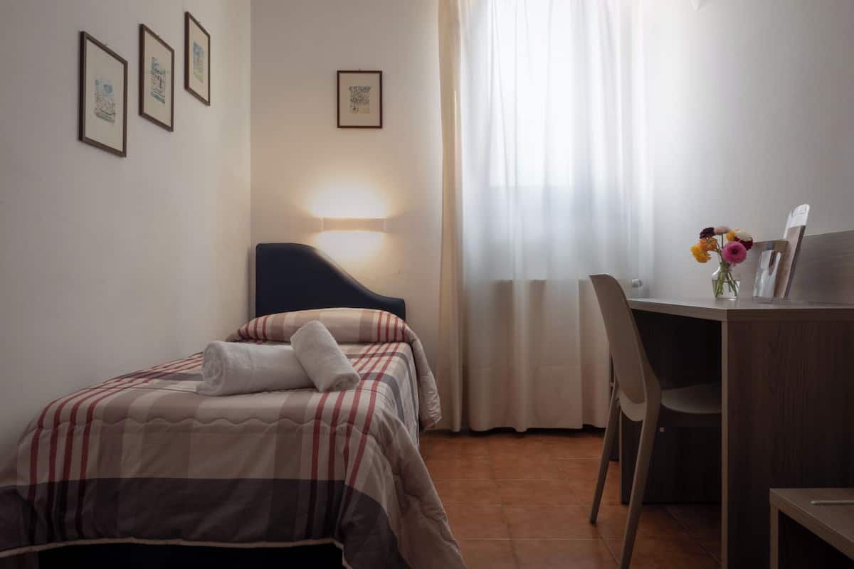 Ein Schlafzimmer mit Bett, Schreibtisch, Stuhl, Lampe und einem fenster mit Vorhängen.