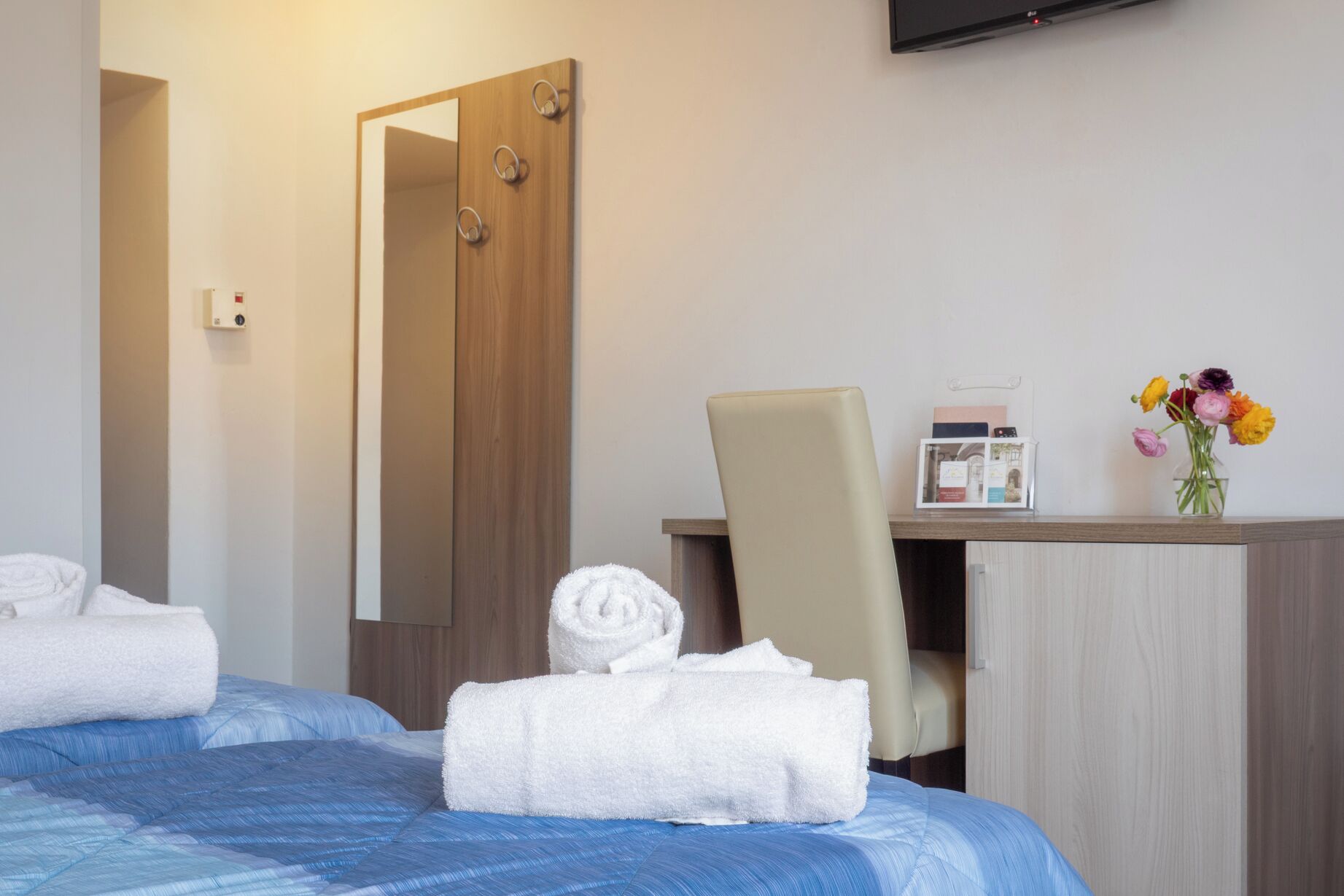 Una habitación de hotel con una cama, un escritorio con silla, un espejo, un televisor y un florero con flores.