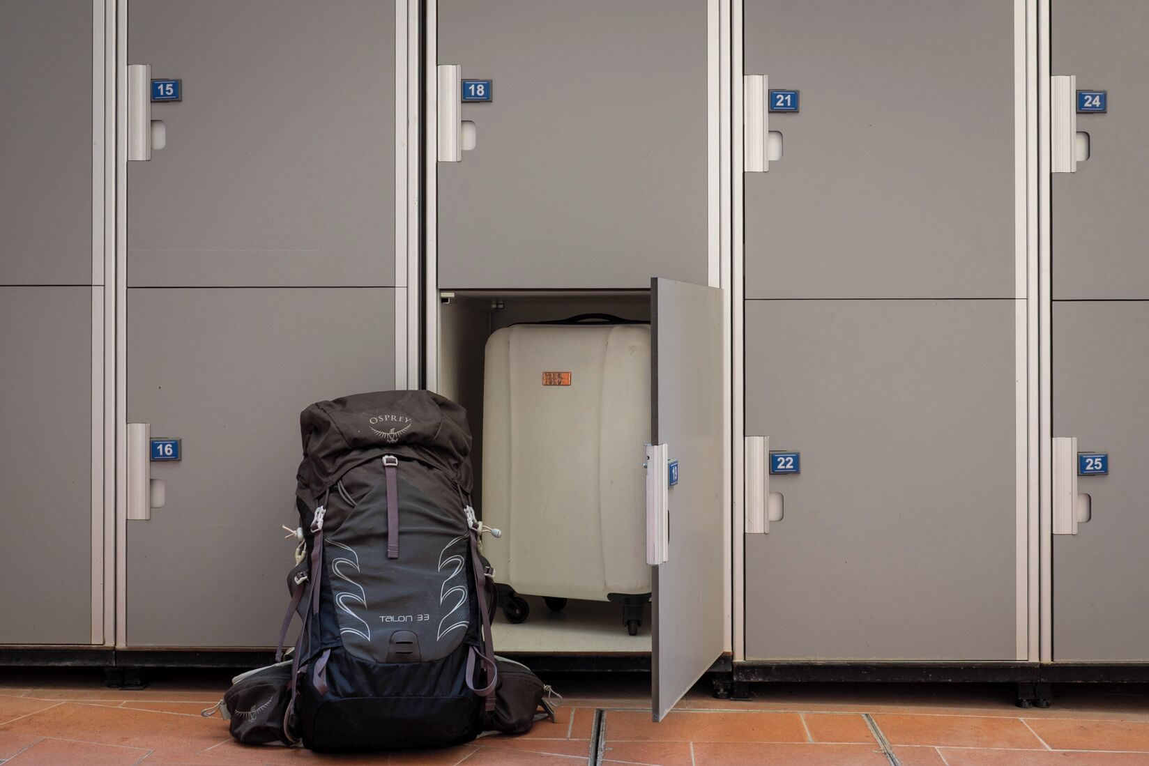 A row of grey lockers with numbers 15 to 25, a black Talon 33 backpack, and a white suitcase in locker 18.