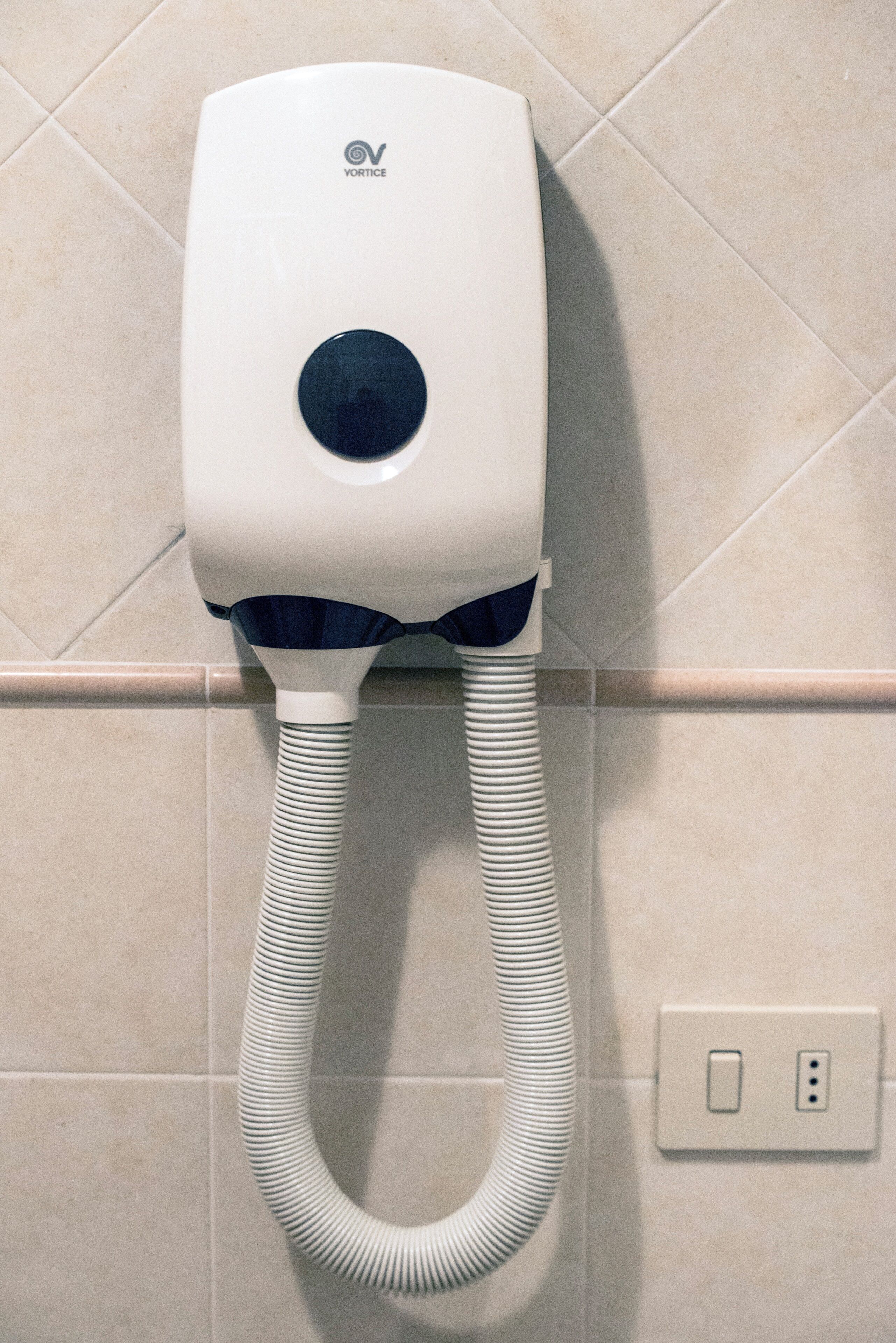 A white hair dryer with a cord, mounted on a tiled wall with a power outlet.