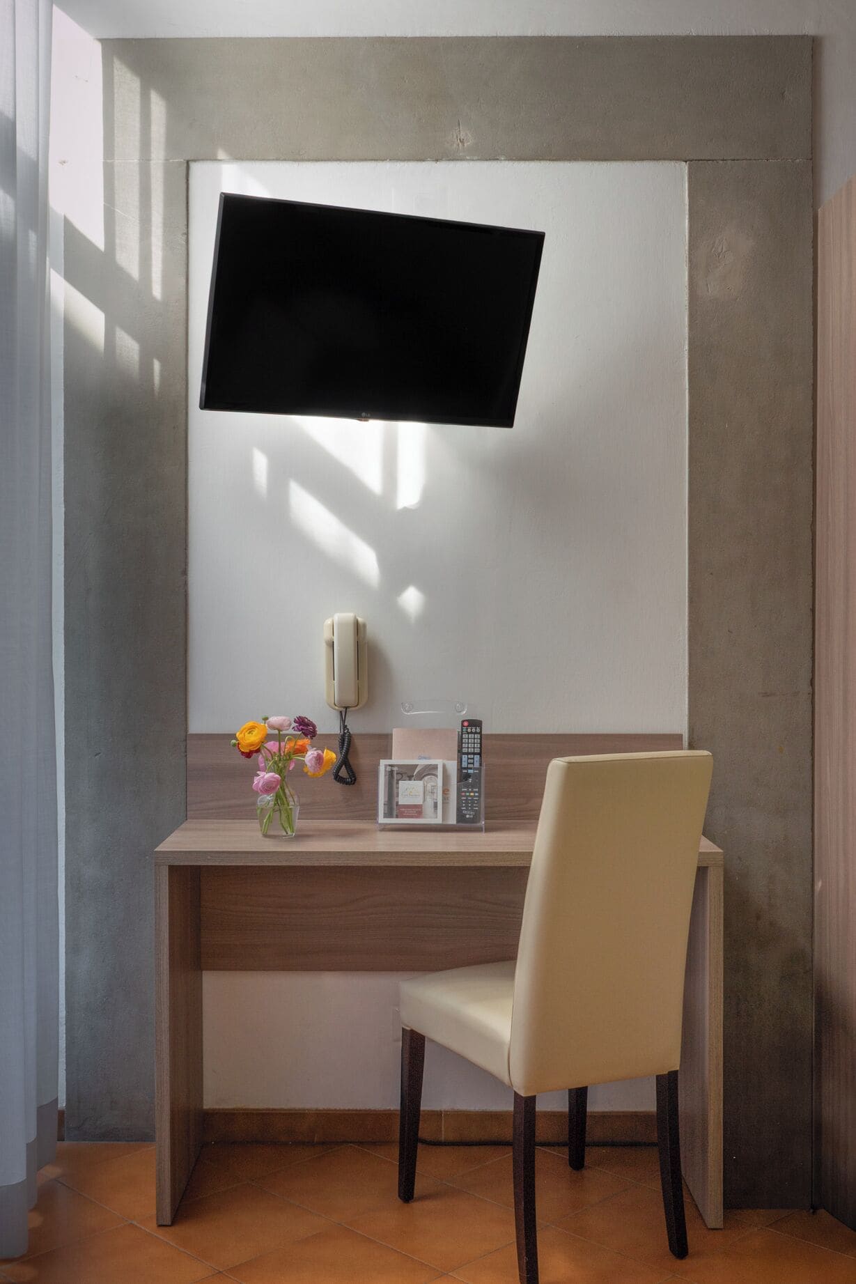 A modern office desk with a mounted flat-screen TV, a telephone, a remote control, and a vase with flowers.