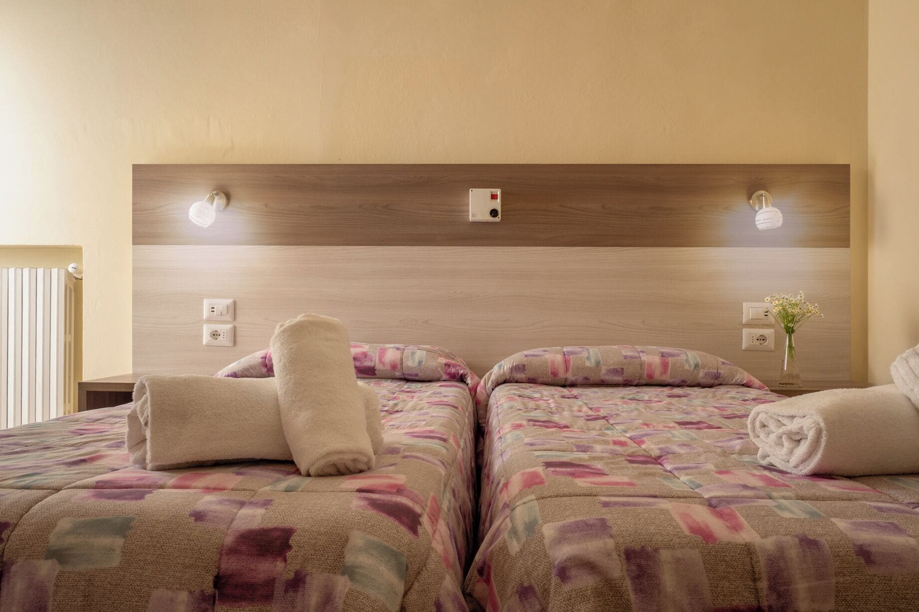 A hotel room with two beds, each with a floral-patterned bedspread, a wooden headboard, bedside lamps, and a vase with flowers on the nightstand.