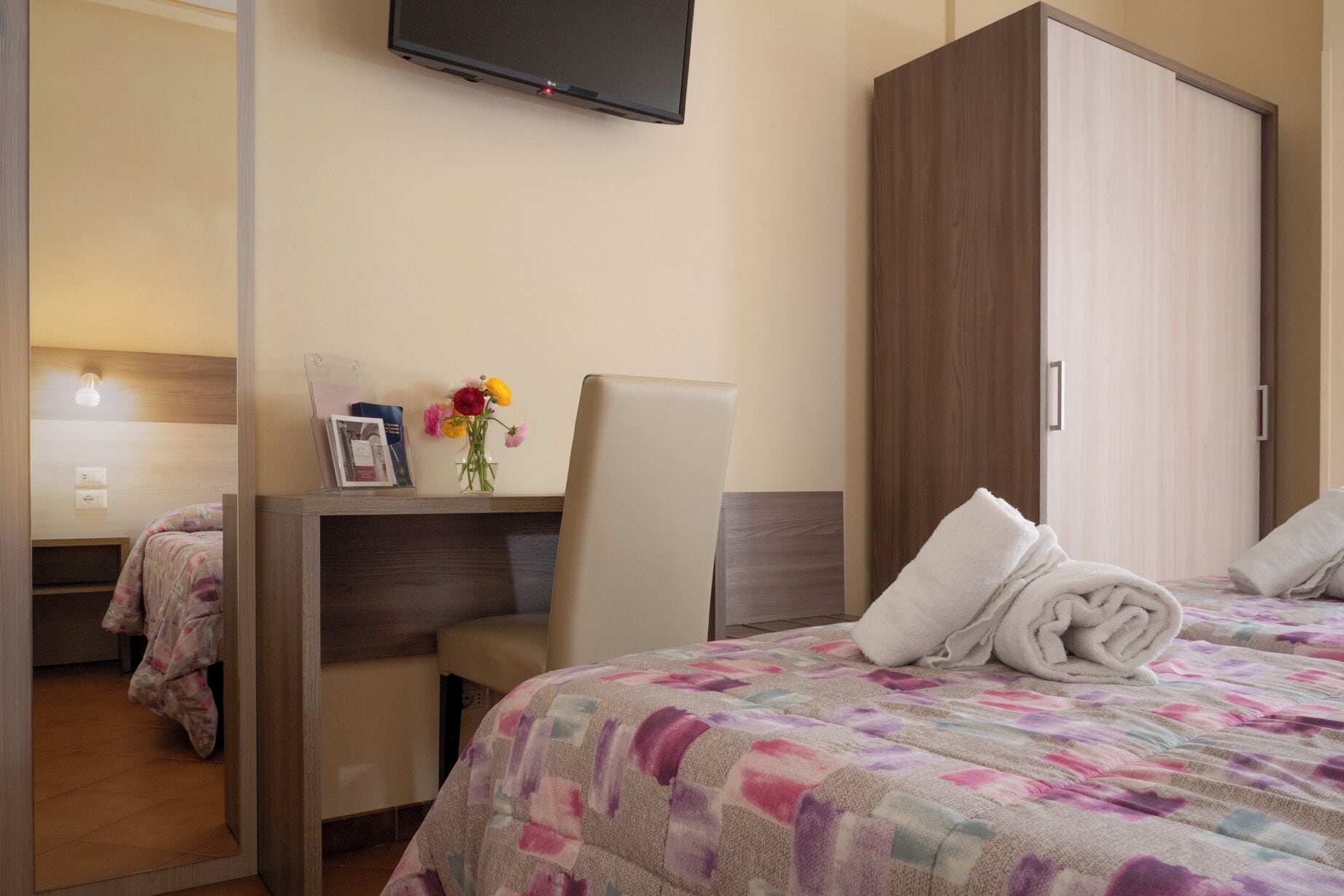 Ein Hotelzimmer mit Bett, Schreibtisch, Stuhl, Kleiderschrank und einem an der Wand befestigten Fernseher.
