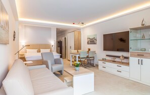 A modern hotel room with a sofa, a dining table with chairs, a TV, and a minibar.