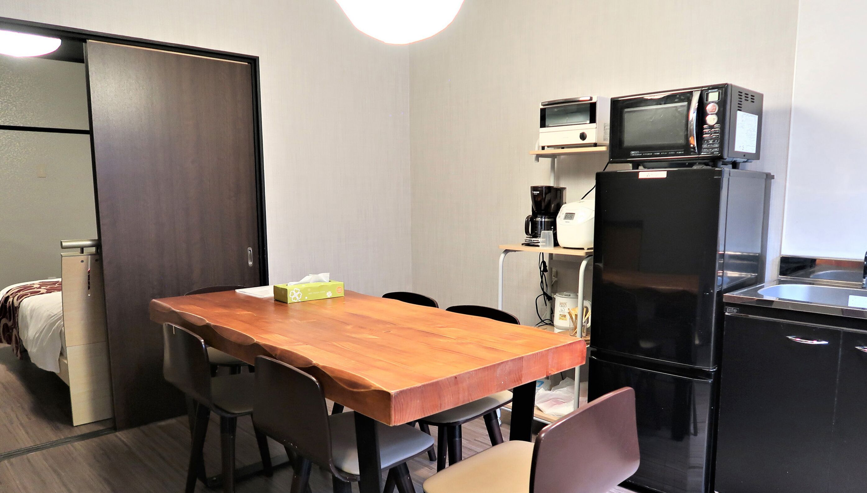 A compact hotel room with a wooden dining table, four chairs, a kitchenette with a microwave, coffee maker, and a refrigerator.