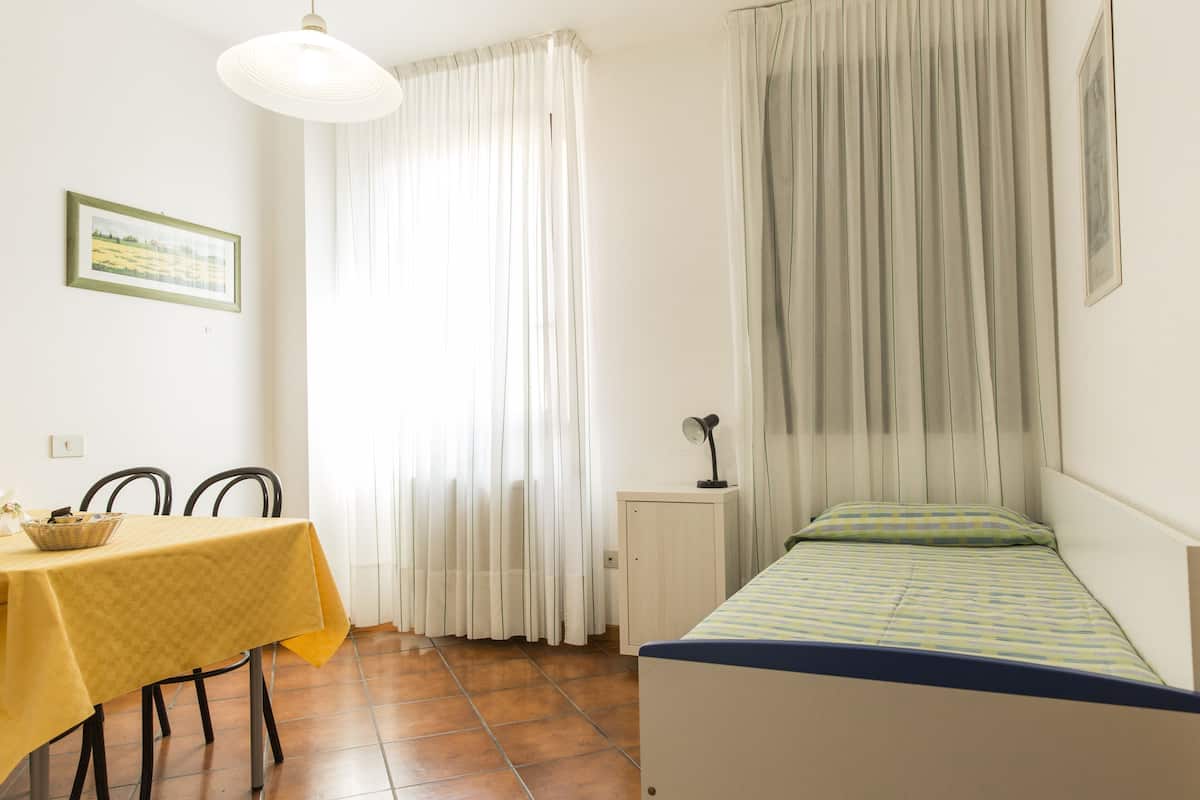 A room with a bed, a table with a yellow tablecloth, a chair, a lamp, and a window with curtains.