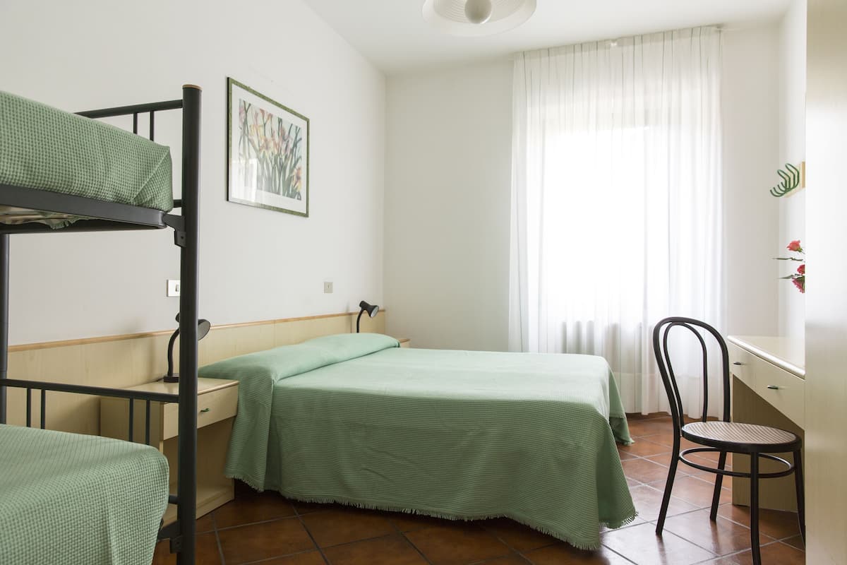 A dormitory room with bunk beds, a desk, a chair, and a window with curtains.