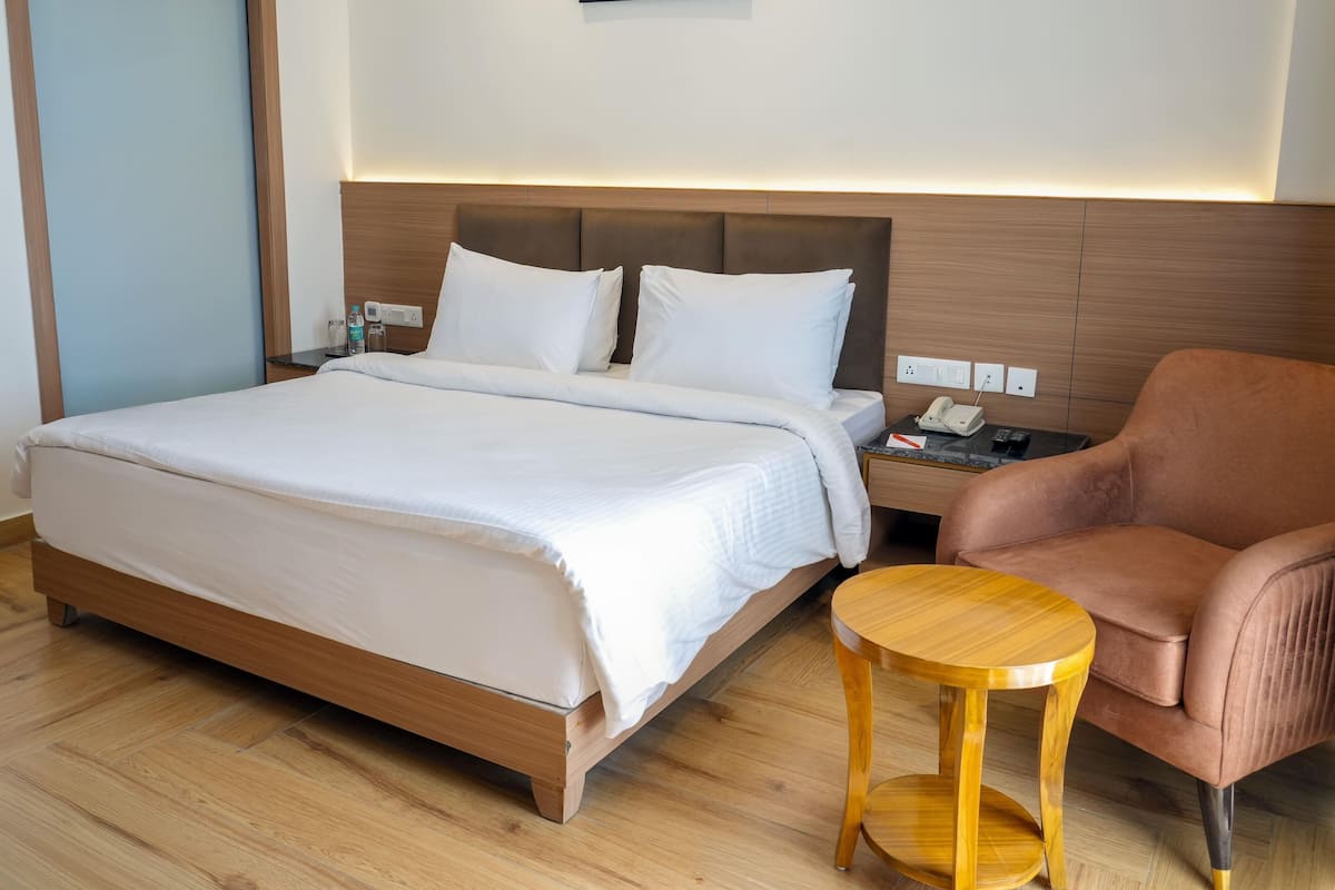 Une chambre d’hôtel avec un grand lit, une table de chevet en bois avec un téléphone et une bouteille, un fauteuil brun et une petite table en bois.