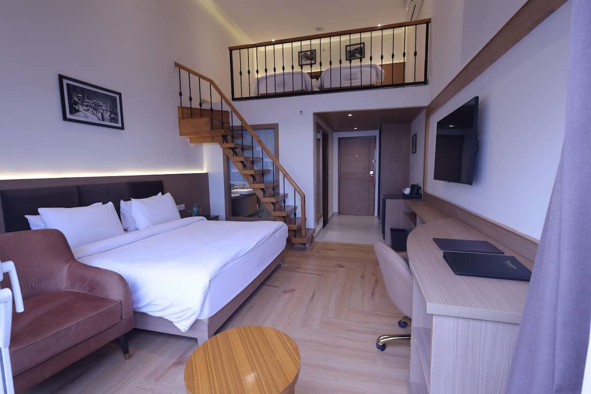 Une chambre d’hôtel moderne avec un grand lit, un canapé en cuir marron, un bureau en bois et un escalier menant à un niveau supérieur.