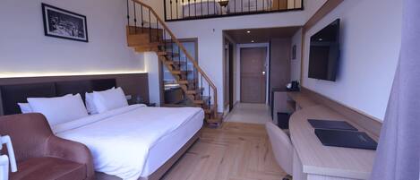 A modern hotel room with a large bed, a brown leather sofa, a wooden desk, and a staircase leading to an upper level.