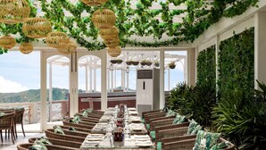 A long dining table set for a meal in a room with wicker chairs, hanging wicker lamps, and lush green foliage on the walls. - Ramada by Wyndham Mussoorie Mall Road (Dehradun)