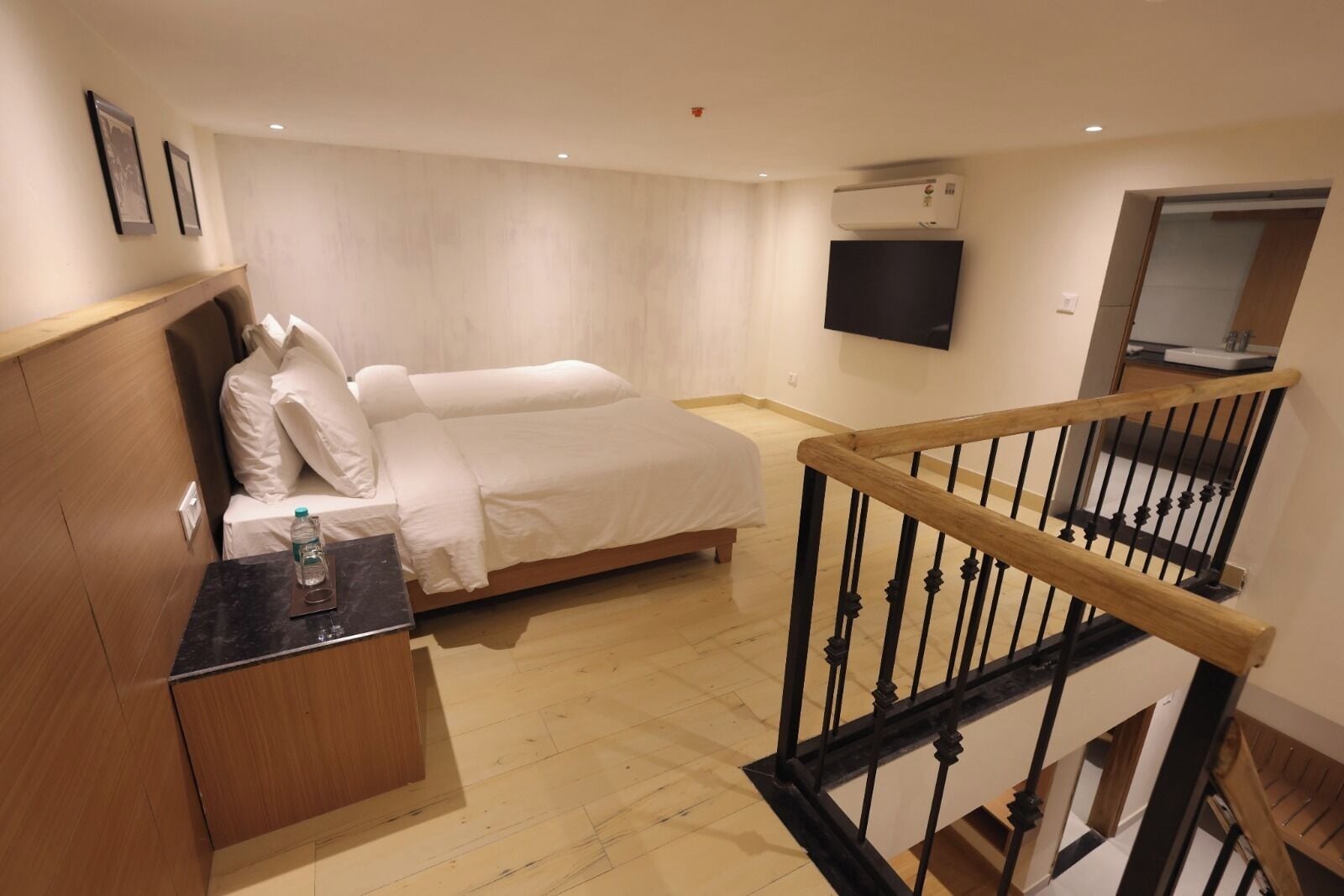A hotel room with a bed, a desk with a water bottle, a TV mounted on the wall, and a wooden staircase.