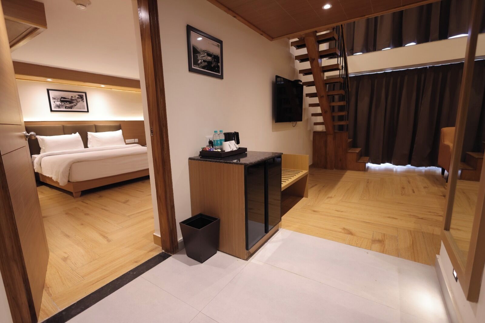 A hotel room with a bed, a desk with a chair, a trash bin, a TV mounted on the wall, and a staircase with a wooden railing.