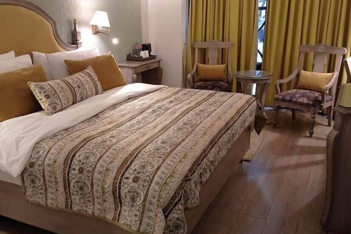 Une chambre d’hôtel avec un grand lit, une table de chevet, une chaise, une petite table et une fenêtre avec des rideaux.