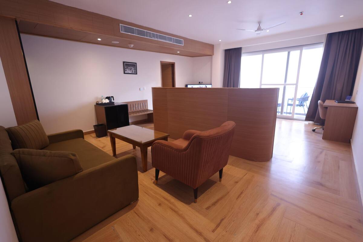 Une chambre d’hôtel moderne avec un plancher en bois, un canapé marron, un fauteuil rouge, une petite table et un bureau avec un ordinateur.