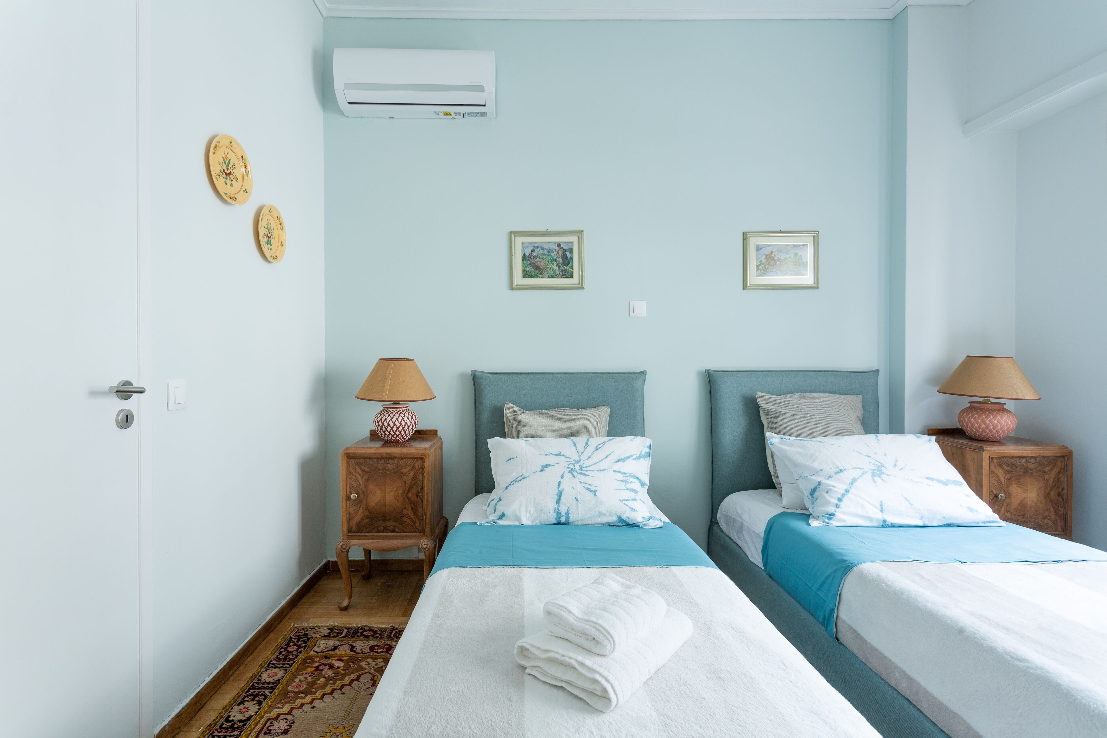 A two-bed room with a light blue wall, a wooden nightstand with a lamp, and a bed with a blue and white bedspread.