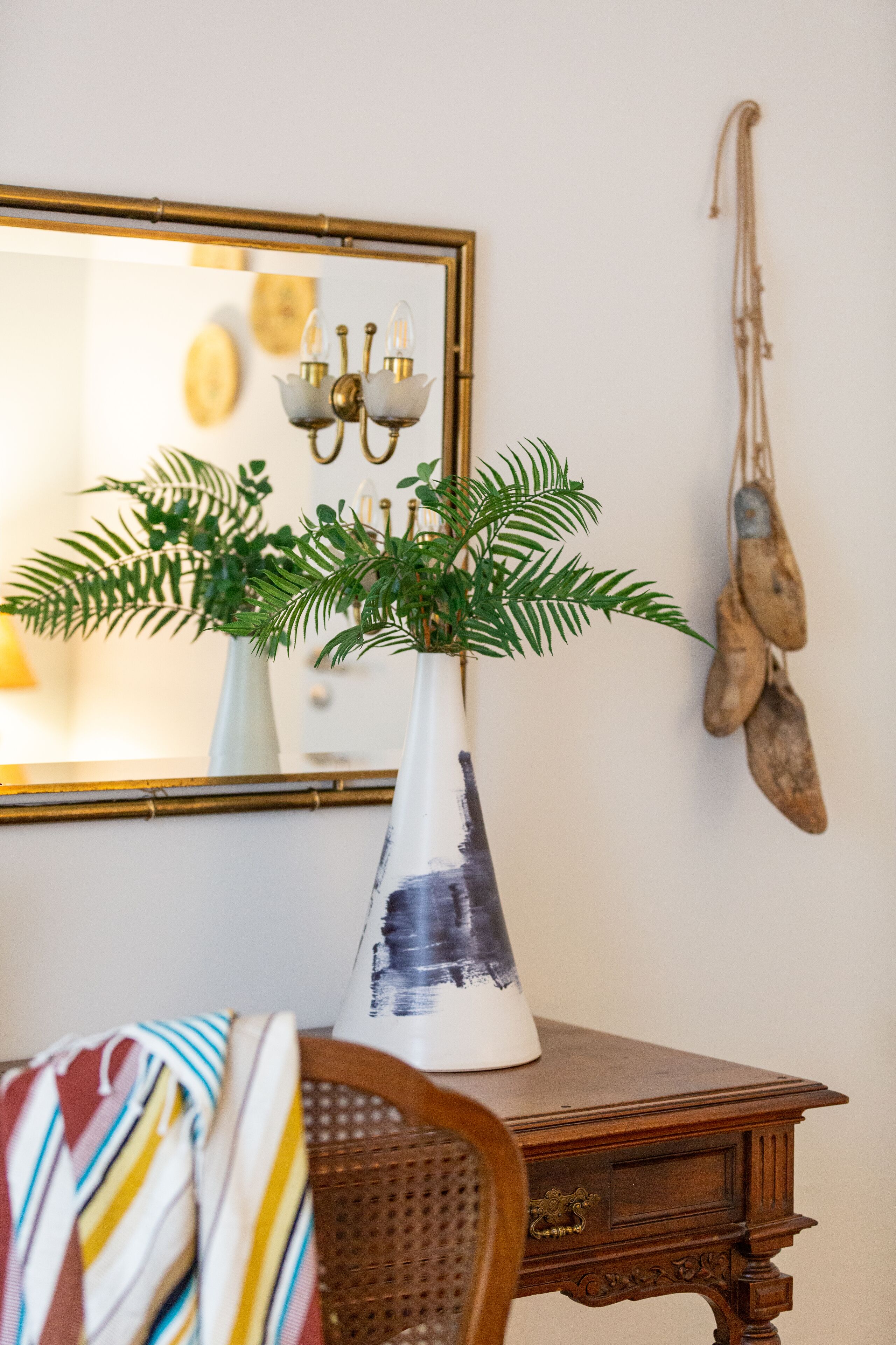 A wooden table with a white vase containing green foliage, a mirror with a gold frame, and a wall-mounted rope swing.